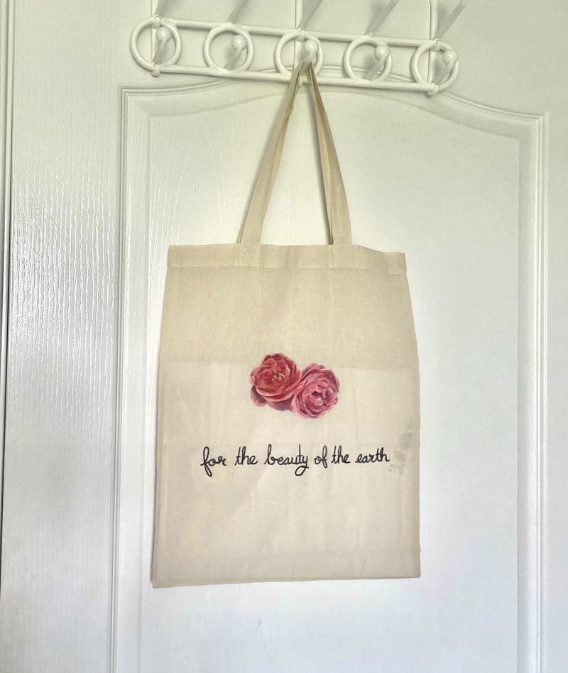Cream cotton tote hand-painted with two full pink roses above the line “for the beauty of the earth” in script.