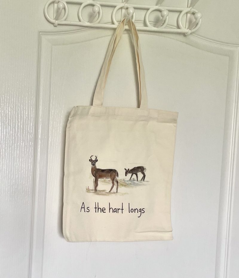 Cream cotton tote hand-painted with a buck and a grazing fawn at a stream above the line “As the hart longs”.