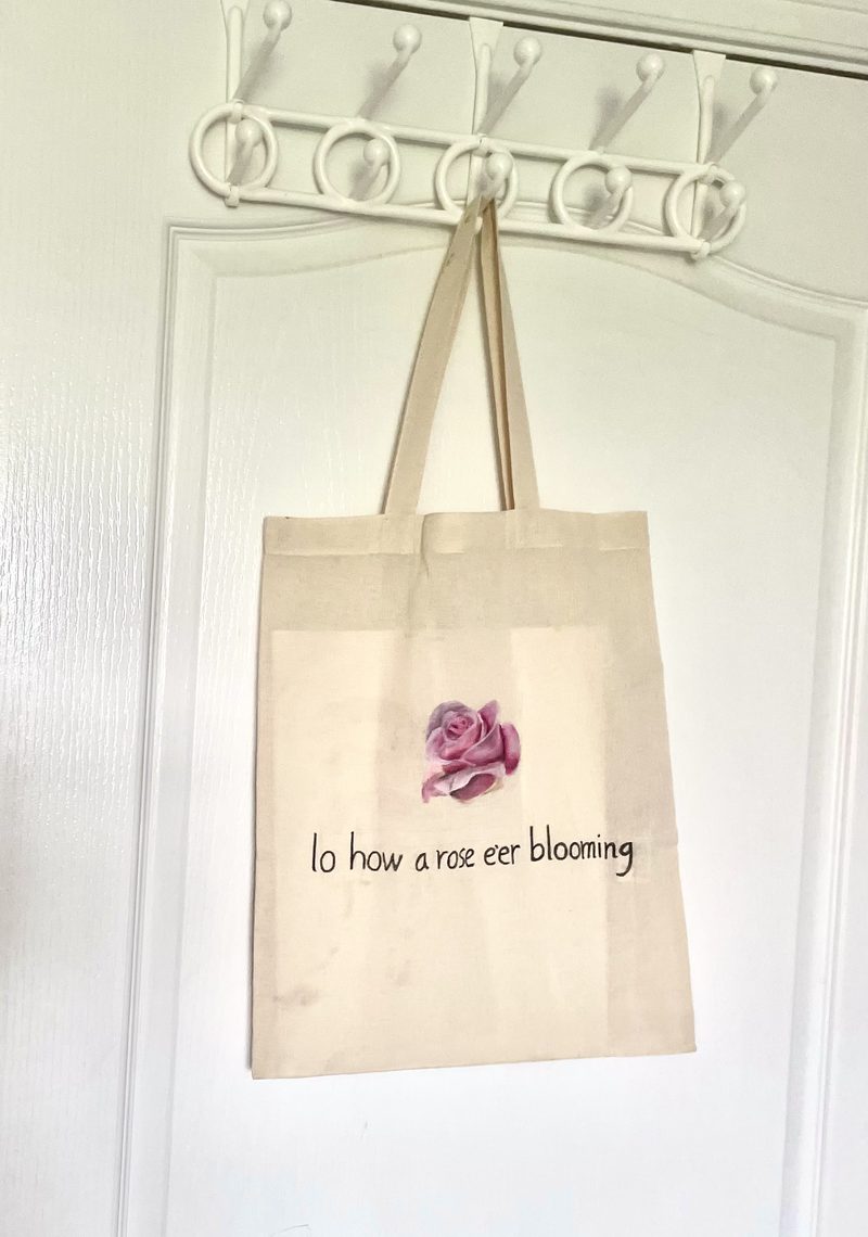 Cream cotton tote hand-painted with a single open pink rose above the line “lo how a rose e’er blooming”.