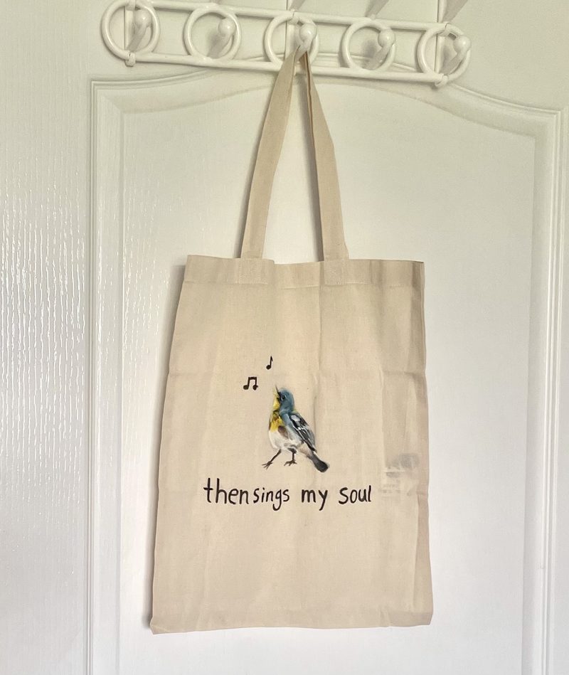 Cream cotton tote hand-painted with a blue and yellow warbler mid-song with two music notes, above the line “then sings my soul”.